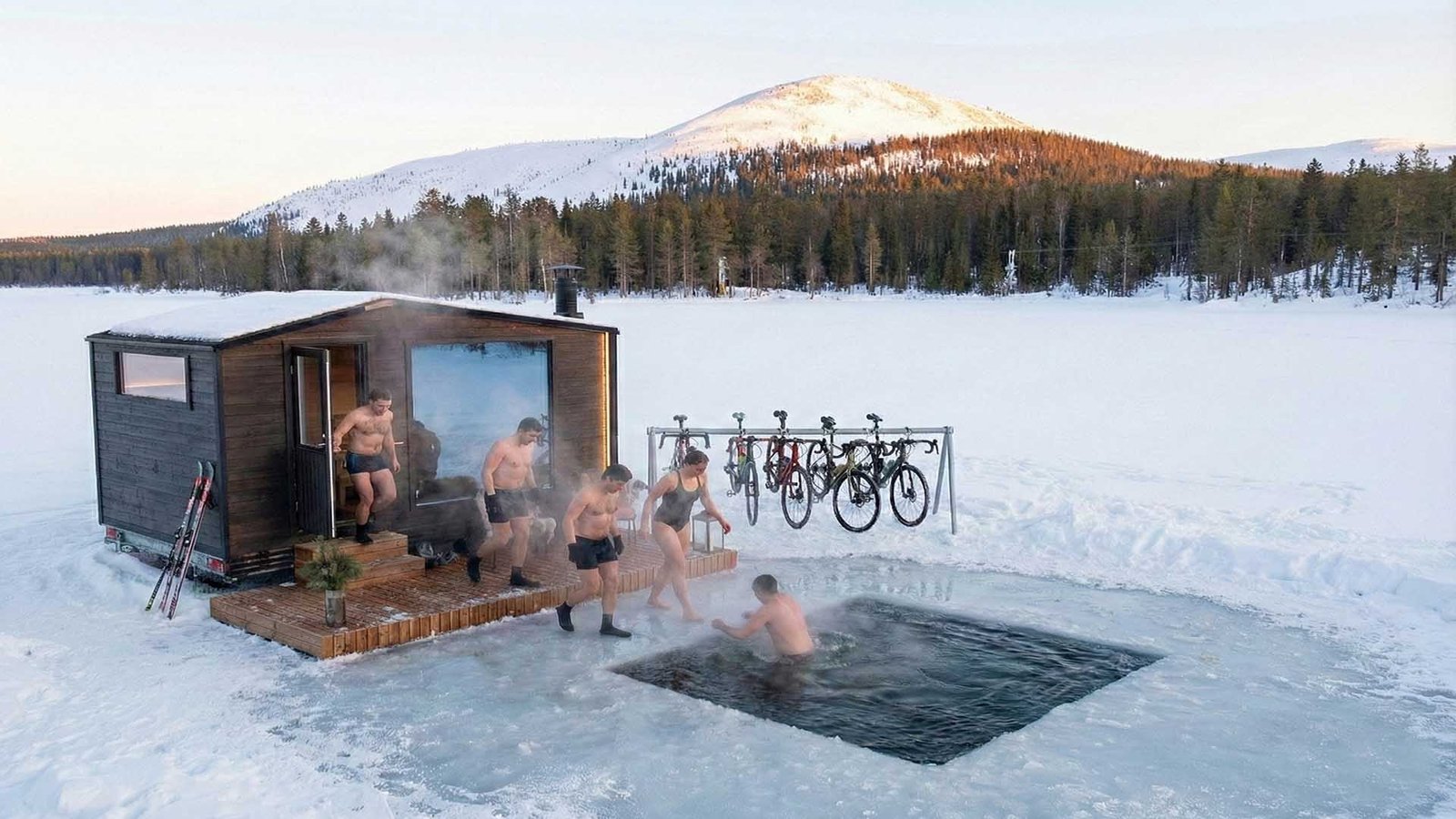 Sauna y baño frío para ciclistas en un lago helado: grupo saliendo de una sauna de leña hacia una inmersión en agua fría con bicicletas al lado.