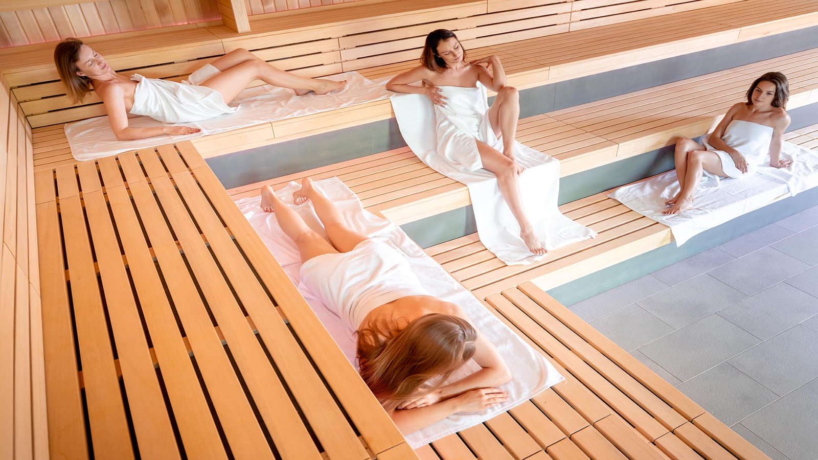 Interior de sauna con bancos de madera y varias personas relajándose sobre toallas.