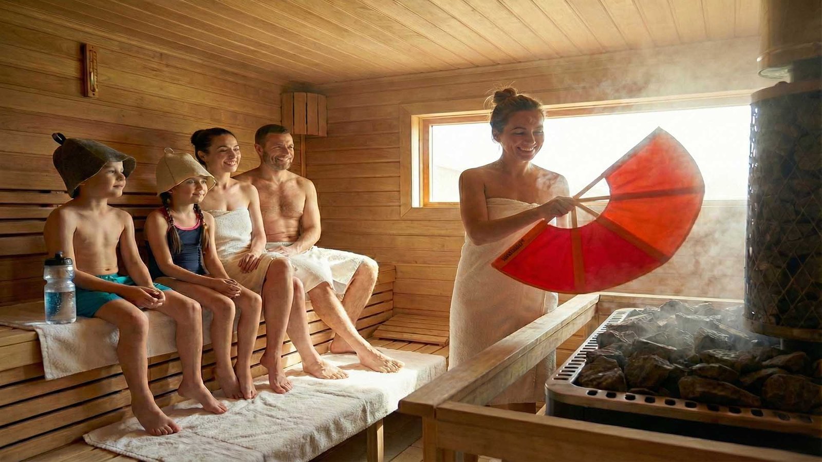 Familia con dos niños en sauna finlandesa mientras la sauna master aviva el vapor con un abanico.